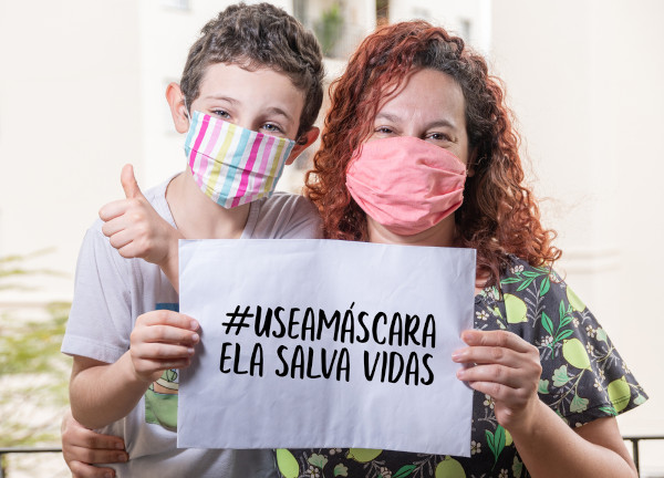 Mulher e garotinho, usando máscaras de proteção facial, seguram juntos placa, em que se lê: “#usemáscara — ela salva vidas”