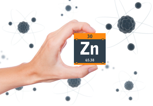 Pessoa segurando um cubo preto com laranja com o símbolo, o número atômico e a massa do elemento químico zinco.