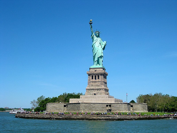 Grande escultura verde de uma mulher em pé erguendo uma tocha, conhecida como a Estátua da Liberdade.