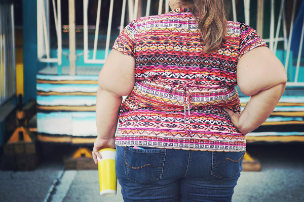 Mulher obesa de costas segurando um copo; a obesidade está associada com alimentos ultraprocessados.