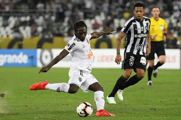 Jogador chutando a bola no campo, em jogo da Copa Sul-Americana; ao fundo, jogador do time adversário e o juiz.