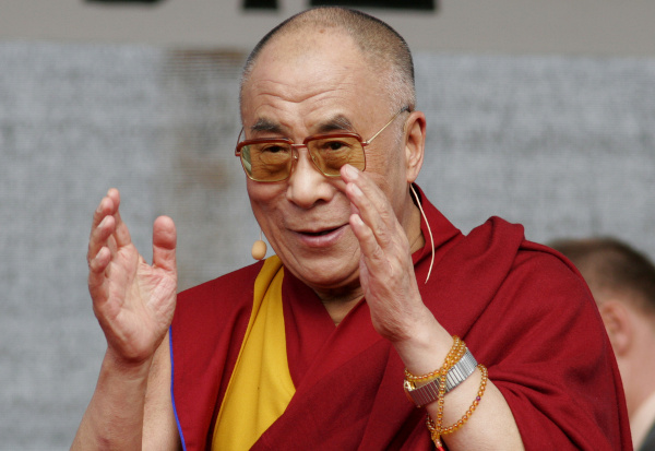 Tenzin Gyatso, o atual Dalai Lama, vestido com roupas típicas e óculos, com as mãos erguidas na altura do rosto.
