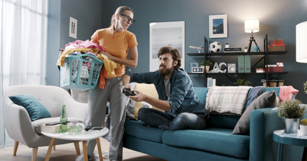 Mulher arrumando a casa enquanto seu marido joga videogame como representação dos estereótipos de gênero.