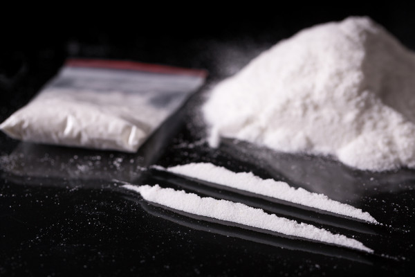 Pacote de plástico com cocaína; ao lado, duas linhas e pilha de cocaína em fundo preto.