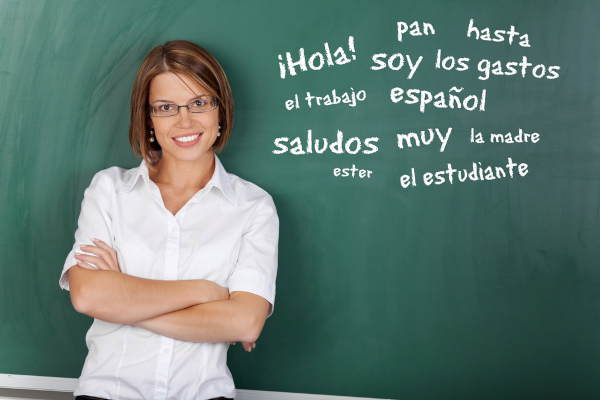 Professora ao lado de um quadro-negro com palavras e expressões de vocabulário estudado pela gramática da língua espanhola.
