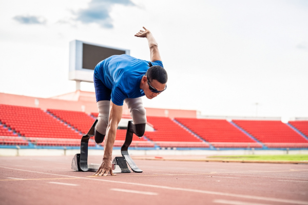 Homem asiático com óculos com duas próteses nas pernas saindo da posição de larga em uma pista de atletismo.