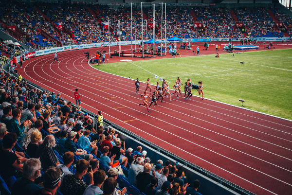 Estádio com público na arquibancada enquanto atletas mulheres realizam corrida na pista de atletismo.