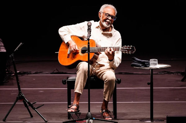 Gilberto Gil sentado tocando violão em apresentação musical.