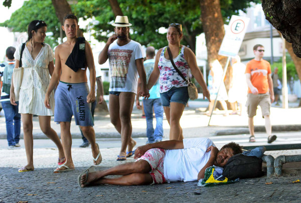 Pedestres observando um homem dormindo na calçada, exemplificando a diferença entre as classes sociais no Brasil.