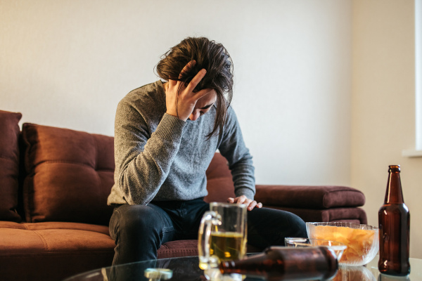 Homem coloca mão na cabeça diante de várias bebidas, em referência ao alcoolismo. [imagem_principal] 