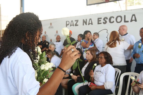 Pessoas na escola segurando rosas brancas, menina negra de costas em primeiro plano. Quadro escrito paz nas escolas