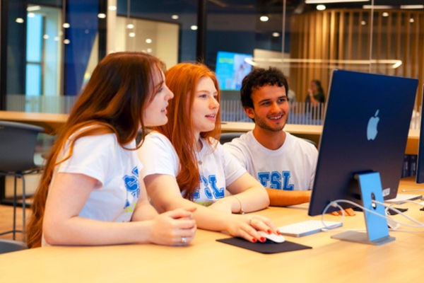 Imagem de três estudantes da Faculdade Sírio-Libanês conversando enquanto olham para a tela de um computador na biblioteca da faculdade.